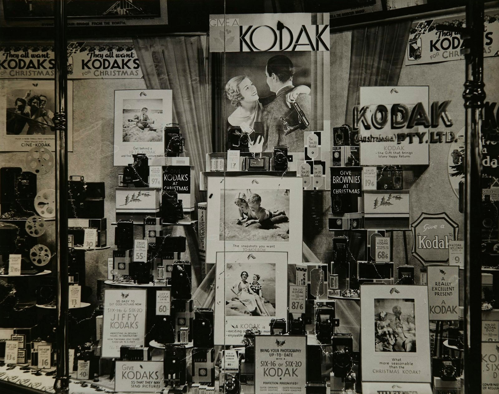 Vitrine antiga em preto e branco de uma loja Kodak com o anúncio Give a Kodak, câmeras e materiais publicitários dos anos 1930